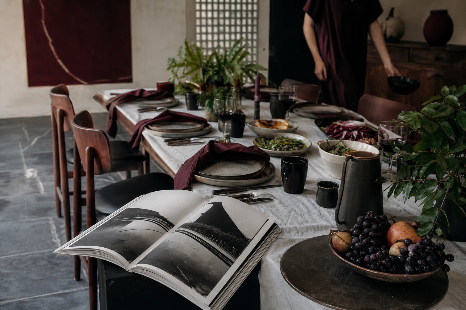 Mesa editorial com cerâmicas, alimentos, folhagens e publicação aberta em um interior modernista.
